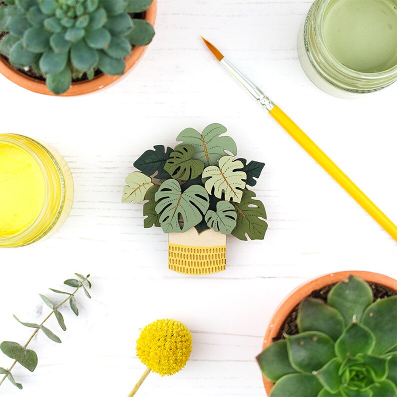 May include: A wooden monstera plant decoration in a yellow pot with a textured design. The leaves are various shades of green. The image also includes a paintbrush, succulents, and paint jars.