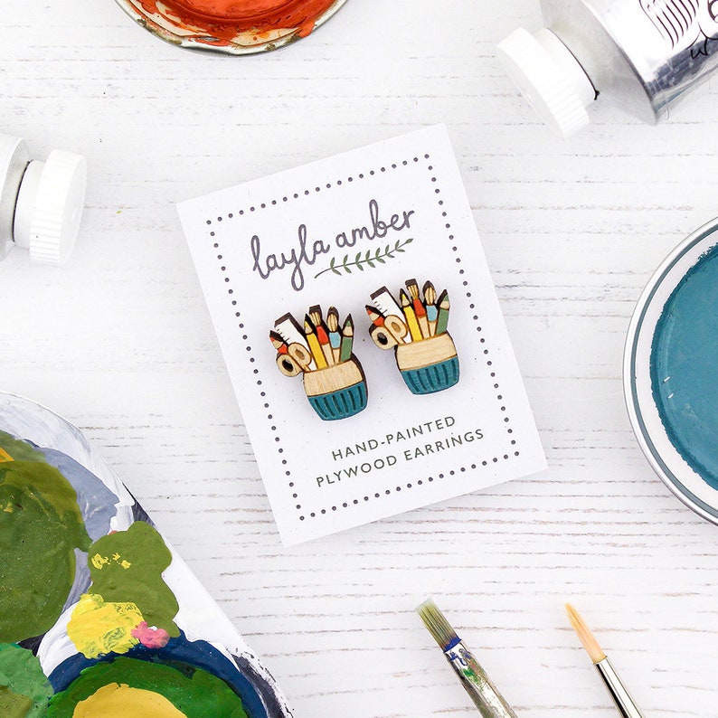 May include: A pair of hand-painted wooden earrings featuring a small, teal-colored cup filled with colorful pencils, crayons, and a ruler. The earrings are displayed on a white card with the text "layla amber" and "HAND-PAINTED PLYWOOD EARRINGS".