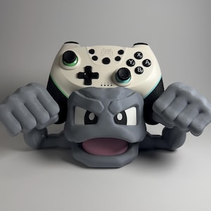 May include: A gray, cartoon-style figure with clenched fists supports a white and black video game controller. The controller has black buttons and green accents. The figure has a wide open mouth and white eyes.