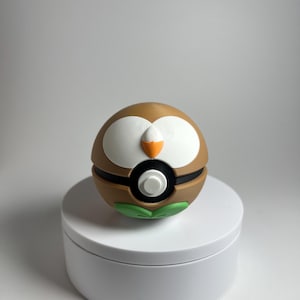 May include: A spherical object designed to resemble a creature with a brown upper half, white eye patches, and an orange beak. It features a black horizontal band across the centre with a white circular button and green leaf-like shapes at the base.