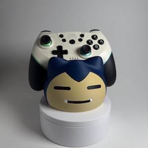 May include: A white and black video game controller with green accents and black buttons. A blue and tan Snorlax-shaped accessory is in front of the controller, resting on a white circular stand. The controller has the letters A, B, X, and Y.
