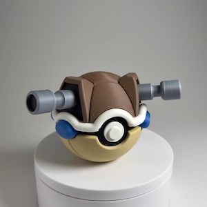 Peut inclure: Une Poké Ball imprimée en 3D, aux couleurs marron, beige, blanc et bleu. Elle est ornée de deux extensions cylindriques grises. Le design est inspiré d'une série animée populaire.