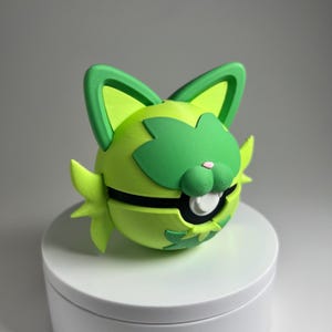 May include: A 3D-printed, lime green and dark green cat-like character with a round body, large ears, and a smiling face. The character has a black band around its middle and a white mouth. The design is whimsical and playful.