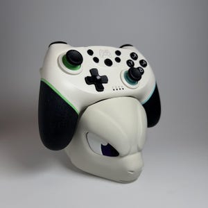 May include: A white and black video game controller with green accents, resting on a light gray Mewtwo-shaped stand. The controller features black buttons and a directional pad. The stand has a detailed face.
