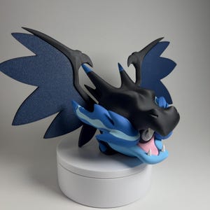 May include: A detailed figurine of a dragon-like creature with a black head and wings, and a blue body. The creature has an open mouth with pink and white details. The wings are a dark blue with a textured appearance. The figurine is displayed on a white platform.