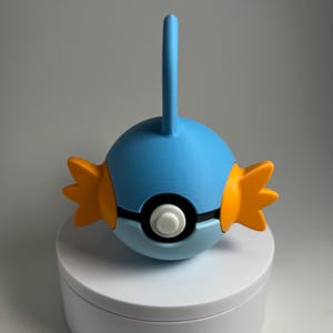 May include: A blue and orange Mudkip figure, a Pokémon character, with a white and black circular design. The figure has a fin on top and orange fins on the sides. The figure is on a white surface.