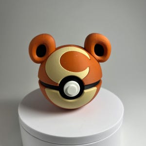May include: A 3D-printed orange and cream-colored ball with a white button and black accents. The ball has two ear-like protrusions and a large "C" design on the front. The object is sitting on a white surface.