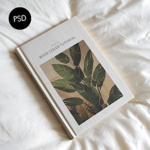 May include: A hardcover book titled "This is a Book Cover Tutorial" with a botanical illustration on the cover. The cover features a close-up of green leaves against a light brown background. The book is lying on a white, textured surface.