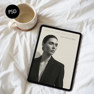 May include: A digital tablet displaying a website mockup with the text "YOUR DESIGN HERE" and a model wearing a black blazer. A beige ceramic mug with a cork base sits beside the tablet on a white sheet.