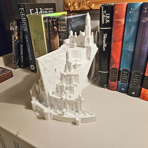 May include: A white, 3D-printed model of a castle, used as a bookend, sits on a white surface. Books of various colors and sizes are arranged behind the bookend. The bookend is approximately 10 inches long.