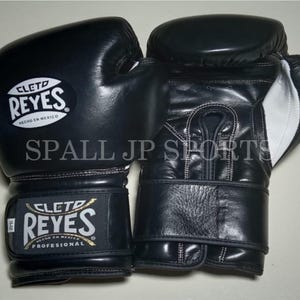 Peut inclure: Paire de gants de boxe en cuir noir avec coutures et accents blancs. Les gants présentent le logo "CLETO REYES" et le texte "HECHO EN MEXICO". Le texte "SPALL JP SPORTS" est également visible.