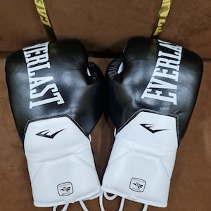 Puede incluir: Un par de guantes de boxeo negros y blancos con el logotipo de Everlast en letras blancas en la parte superior. La zona de la muñeca es blanca con un pequeño logotipo y el texto Hecho en Mexico impreso en negro. Cordones blancos visibles.