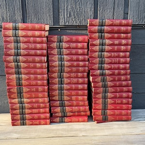 Puede incluir: Montones de libros antiguos con cubiertas rojas y letras doradas. Los libros están dispuestos en tres columnas, con los lomos orientados hacia el espectador. Los títulos están impresos en dorado sobre una banda negra. Los libros están sobre una superficie de madera.