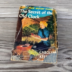 May include: Vintage Nancy Drew Mystery Stories book, "The Secret of the Old Clock," with a cover illustration of a woman reading. The book's cover is worn, showing signs of age and use. The author's name, Carolyn Keene, is visible.