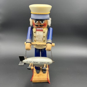 May include: A wooden nutcracker in the form of a military officer, wearing a white and blue uniform and a hat. The nutcracker holds a small silver zeppelin model with the text "LZ 127 GRAF ZEPPELIN". The word "ONSTRUKTIONE" is written on a wooden bar.