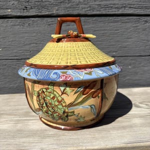 May include: A decorative ceramic jar with a lid. The jar features a turtle design with green, brown, and tan colors. The lid is yellow with a brown handle. The jar has a blue and floral design around the rim.
