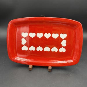 Puede incluir: Una bandeja rectangular de cerámica con un acabado rojo brillante. El centro de la bandeja presenta un patrón decorativo compuesto por doce corazones blancos dispuestos en una formación rectangular sobre el fondo rojo sólido.