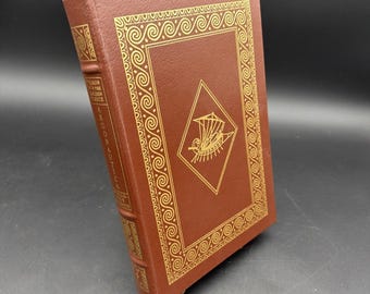 Puede incluir: Un libro de tapa dura marrón titulado Argonautica de Apolonio de Rodas. La portada presenta un marco de diamante estampado en oro con una ilustración de un barco y un borde griego. El lomo muestra el título y el autor en letras doradas.
