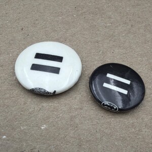 May include: Two circular buttons, one white and one black, each displaying a horizontal equal sign. The white button has a black equal sign, and the black button has a white equal sign. Both buttons have a shiny surface.