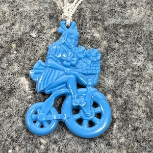 May include: A blue ceramic pendant necklace featuring a girl riding a bicycle with a basket of flowers.