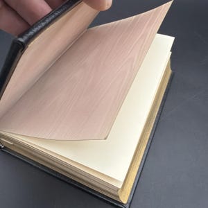 Puede incluir: Un diario encuadernado en cuero negro con bordes dorados. Las p&aacute;ginas son de color crema y la cubierta interior tiene un dise&ntilde;o de vetas de madera. El diario est&aacute; abierto, con una mano pasando una p&aacute;gina, revelando el interior.