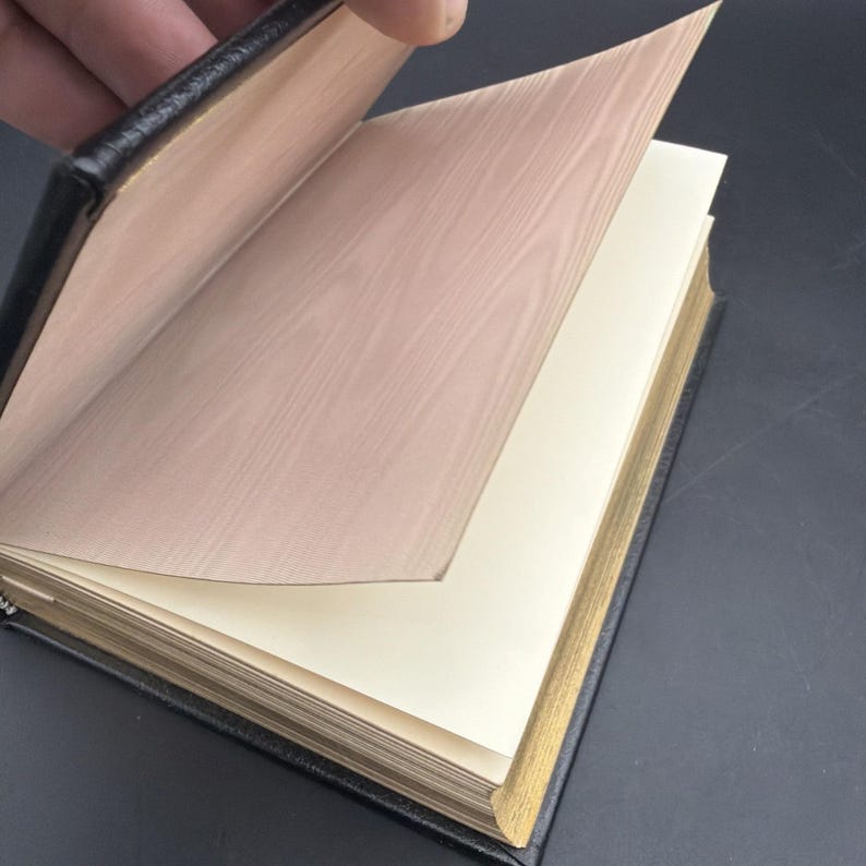 Puede incluir: Un diario encuadernado en cuero negro con bordes dorados. Las p&aacute;ginas son de color crema y la cubierta interior tiene un dise&ntilde;o de vetas de madera. El diario est&aacute; abierto, con una mano pasando una p&aacute;gina, revelando el interior.