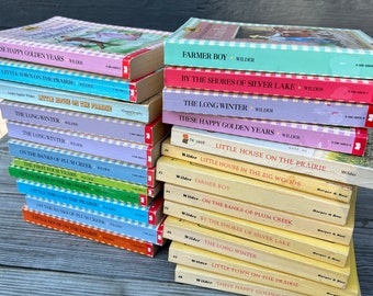 May include: A stack of Little House on the Prairie books by Laura Ingalls Wilder. The books are in various colors, including yellow, blue, red, and green. The titles of the books are visible, including "These Happy Golden Years", "Little House on the Prairie", "Farmer Boy", and "By the Shores of Silver Lake".