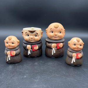 Puede incluir: Cuatro figuras de monjes de cerámica. Cada figura lleva una túnica marrón y sostiene un libro rojo. Los monjes tienen caras sonrientes y son de diferentes tamaños. Una de las figuras es una maceta.