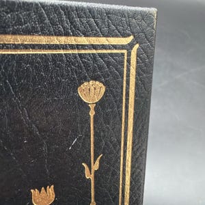Puede incluir: Primer plano de una cubierta de libro en cuero negro con detalles florales dorados. El dise&ntilde;o presenta un borde dorado y una flor dorada estilizada. El cuero tiene un aspecto texturizado.