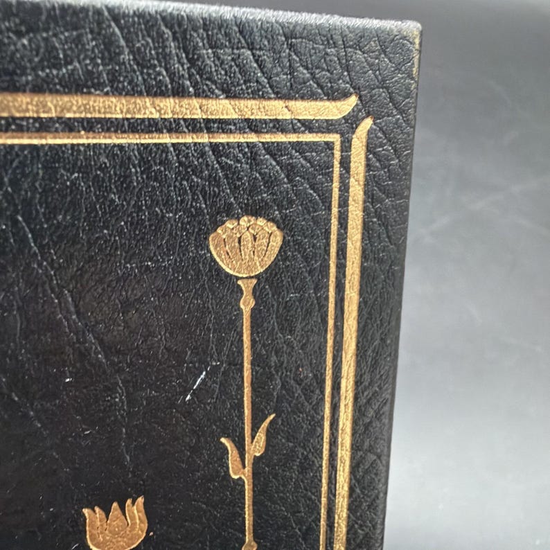 Puede incluir: Primer plano de una cubierta de libro en cuero negro con detalles florales dorados. El dise&ntilde;o presenta un borde dorado y una flor dorada estilizada. El cuero tiene un aspecto texturizado.