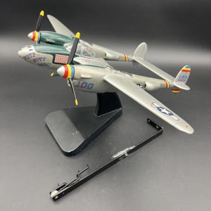 May include: A silver and green P-38 Lightning model aeroplane with twin propellers, featuring the text "PUTT PUTT MARU" and the number "100" on the fuselage. The model is mounted on a black stand, with an additional black metal component nearby.