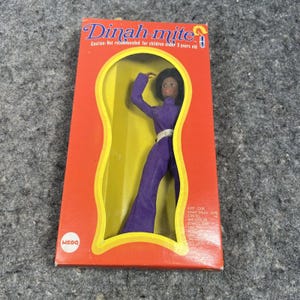 Puede incluir: Una muñeca vintage "Dinah-mite" con un mono morado, exhibida en una caja roja y amarilla. La caja tiene el texto "Dinah-mite" y "Precaución: No recomendado para niños menores de 3 años". La muñeca está en una ventana en forma de ojo de cerradura.