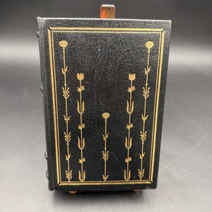 Puede incluir: Un libro negro encuadernado en cuero con dise&ntilde;os florales dorados. La portada presenta un borde rectangular dorado y patrones florales verticales. El libro se exhibe sobre un soporte de madera, mostrando su cubierta y lomo decorativos.
