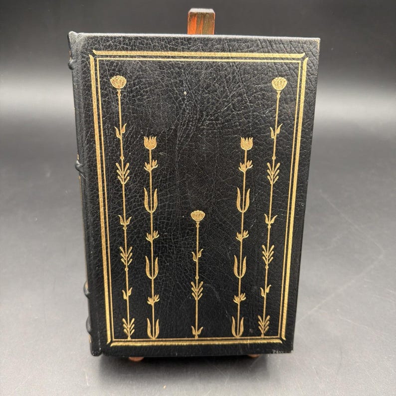 Puede incluir: Un libro negro encuadernado en cuero con dise&ntilde;os florales dorados. La portada presenta un borde rectangular dorado y patrones florales verticales. El libro se exhibe sobre un soporte de madera, mostrando su cubierta y lomo decorativos.