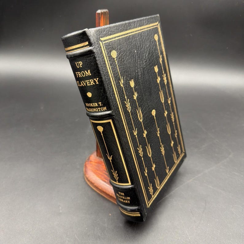 Puede incluir: Un libro de tapa dura negro titulado "Up From Slavery" de Booker T. Washington, con letras doradas y dise&ntilde;os florales, exhibido en un soporte de madera. Se ve el logotipo de The Franklin Library.