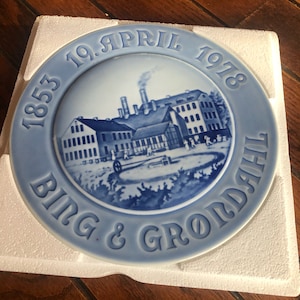 Puede incluir: Un plato de cerámica azul y blanco con una escena de un edificio de fábrica y el texto "1853 19. ABRIL 1978 BING & GRØNDAHL".