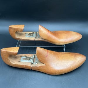 May include: A pair of wooden shoe forms, possibly vintage, are shown. They are light brown with a smooth finish and have a hinged metal mechanism. The forms are on a clear acrylic stand against a dark background.