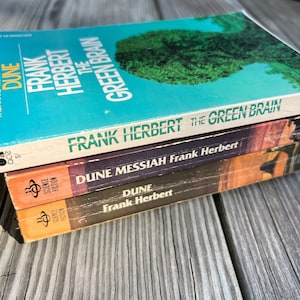 May include: A stack of three paperback science fiction books by Frank Herbert. The top book is "Dune: The Green Brain", the middle book is "Dune Messiah", and the bottom book is "Dune".