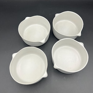 Può includere: Quattro ciotole in ceramica bianca, di forma arrotondata, con un piccolo manico su un lato. Le ciotole sono disposte a quadrato su uno sfondo scuro. Le ciotole sembrano nuove e inutilizzate.