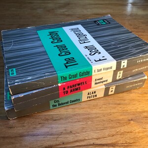 May include: A stack of three paperback books with gray covers and colorful spines. The top book is "The Great Gatsby" by F. Scott Fitzgerald. The middle book is "A Farewell to Arms" by Ernest Hemingway. The bottom book is "Cry, the Beloved Country" by Alan Paton.