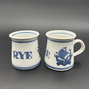 Puede incluir: Dos tazas de cerámica blanca con detalles azules. Una taza tiene la palabra "RYE" en azul, la otra un diseño de velero azul. Ambas tazas tienen borde y base azules, y un asa blanca.