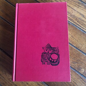 May include: A red hardcover book with a black illustration of a person in a hat playing a drum. The book is on a wooden surface.