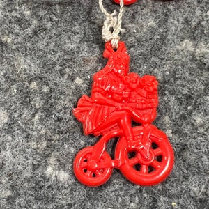 May include: A red plastic charm of a girl riding a bicycle with a basket full of flowers. The charm is attached to a white string.