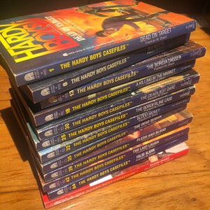 Puede incluir: Una pila de 14 libros de Hardy Boys Casefiles con lomos azules y rojos. Los libros están numerados del 1 al 94, incluyendo los números 8, 12, 18, 25, 30, 32, 35, 39, 42, 49, 54 y 84. Los títulos de los libros son visibles en los lomos, incluyendo "Dead on Target", "See No Evil", "The Borgia Dagger", "A Killing in the Market", "The Deadliest Dare", "The Borderline Case", "Blood Money", "The Dead Season", "Flesh and Blood", "The Last Laugh", "False Alarm", y "Blood Sport".