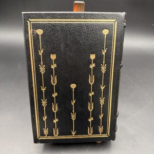 Puede incluir: Caja negra con encuadernaci&oacute;n de cuero y dise&ntilde;o floral dorado. La caja tiene forma rectangular con tapa con bisagras. El dise&ntilde;o floral dorado es sim&eacute;trico y presenta flores y tallos estilizados. Probablemente vintage o antiguo.