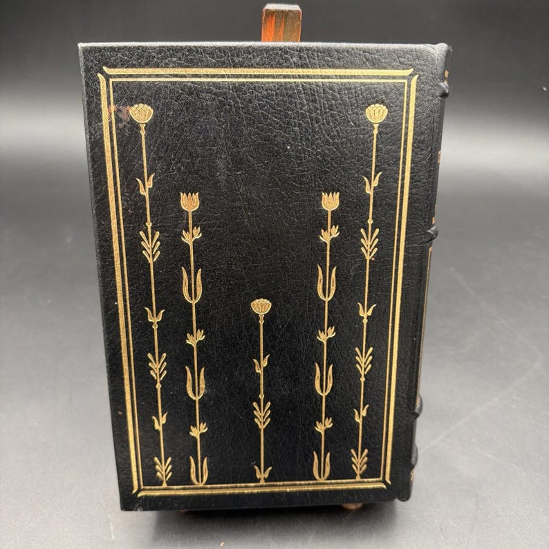 Puede incluir: Caja negra con encuadernaci&oacute;n de cuero y dise&ntilde;o floral dorado. La caja tiene forma rectangular con tapa con bisagras. El dise&ntilde;o floral dorado es sim&eacute;trico y presenta flores y tallos estilizados. Probablemente vintage o antiguo.