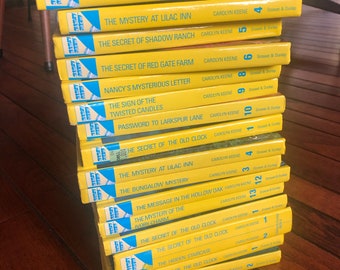 May include: A stack of 16 yellow paperback books with blue and white covers. The books are Nancy Drew mystery novels. The titles of the books are visible, including "The Bungalow Mystery", "The Mystery at Lilac Inn", "The Secret of Shadow Ranch", "The Secret of Red Gate Farm", "Nancy's Mysterious Letter", "The Sign of the Twisted Candles", "Password to Larkspur Lane", "The Secret of the Old Clock", "The Mystery at Lilac Inn", "The Bungalow Mystery", "The Message in the Hollow Oak", "The Mystery of the Ivory Charm", "The Secret of the Old Clock", "The Secret of the Old Clock", "The Hidden Staircase", "The Secret of the Old Clock", and "The Hidden Staircase".