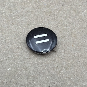 May include: A circular black badge featuring two parallel white lines. The badge has a pin on the back and a small, circular design at the bottom. It rests on a textured, tan surface.