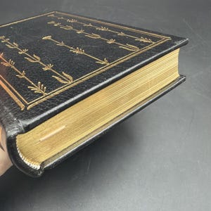 Puede incluir: Un libro de tapa dura con una cubierta de cuero negro y bordes dorados. La cubierta presenta un dise&ntilde;o floral dorado en los bordes. Las p&aacute;ginas son doradas y el libro parece estar en buen estado.