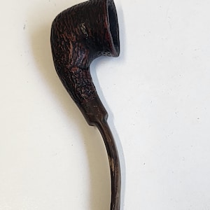 May include: A dark brown tobacco pipe with a deeply textured, rustic bowl and a smooth, slightly curved stem. The bowl features a rugged, pitted surface finish, while the mouthpiece is slender and tapers toward the end, showing a consistent dark wood-toned appearance throughout the entire object.